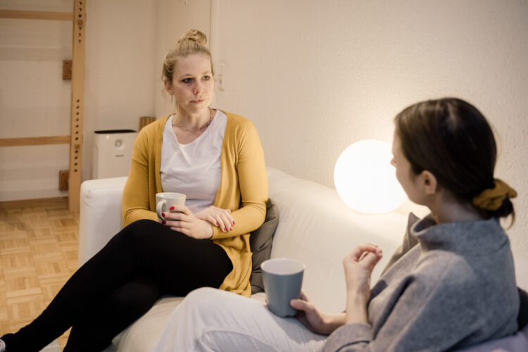 Zwei Frauen sitzen auf einem Sofa mit Tassen in den Händen. Der Raum ist hell erleuchtet und die zwei befinden sich in einem Gespräch.