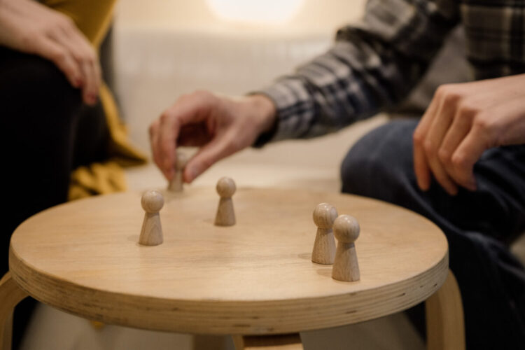 Auf einem Tisch sind mehrere Spielfiguren aus Holz aufgestellt, die von zwei Menschen mit den Händen bewegt werden.